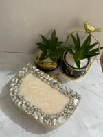 A cream-colored clutch adorned with clear crystals, mother-of-pearl accents, and pearls, placed next to decorative plants.