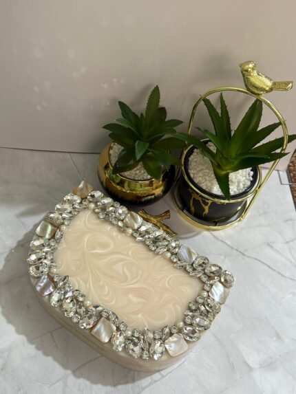 A cream-colored clutch adorned with clear crystals, mother-of-pearl accents, and pearls, placed next to decorative plants.