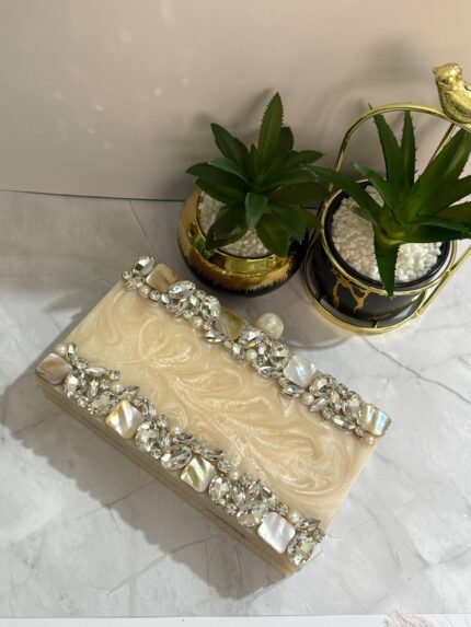 A cream-colored clutch with clear crystals, mother-of-pearl accents, and pearls, placed beside decorative plants.