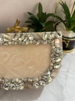 A cream-colored clutch adorned with clear crystals, mother-of-pearl accents, and pearls, placed next to decorative plants.