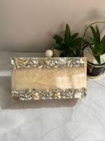A cream-colored clutch with clear crystals, mother-of-pearl accents, and pearls, placed beside decorative plants.