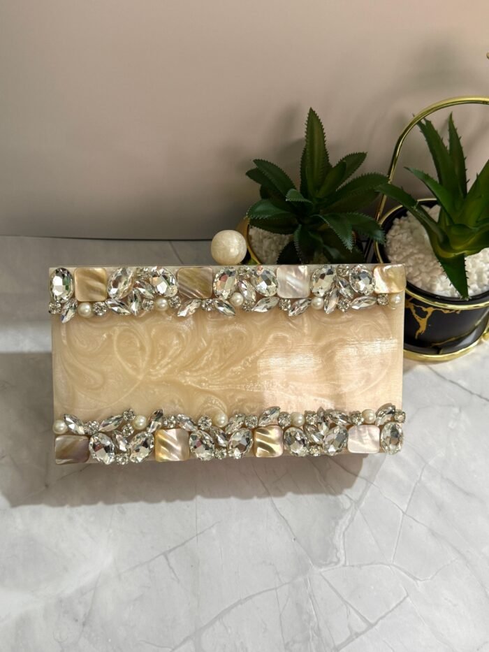 A cream-colored clutch with clear crystals, mother-of-pearl accents, and pearls, placed beside decorative plants.