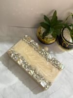 A cream-colored clutch with clear crystals, mother-of-pearl accents, and pearls, placed beside decorative plants.