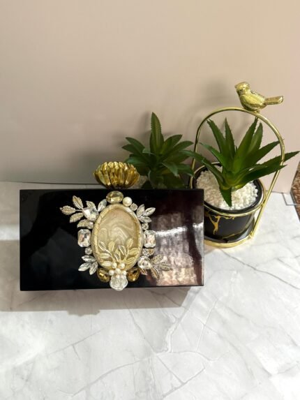 A black clutch with floral and gemstone accents, featuring a central mother-of-pearl design, gold-toned leaves, and pearls, placed next to decorative plants.