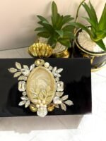 A black clutch with floral and gemstone accents, featuring a central mother-of-pearl design, gold-toned leaves, and pearls, placed next to decorative plants.