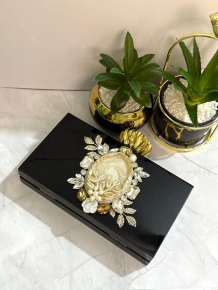 A black clutch with floral and gemstone accents, featuring a central mother-of-pearl design, gold-toned leaves, and pearls, placed next to decorative plants.