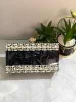 A black clutch adorned with a border of large clear crystals, placed next to decorative plants and a ring.