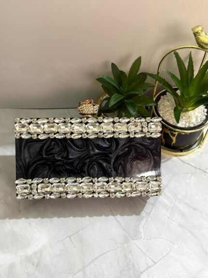 A black clutch adorned with a border of large clear crystals, placed next to decorative plants and a ring.