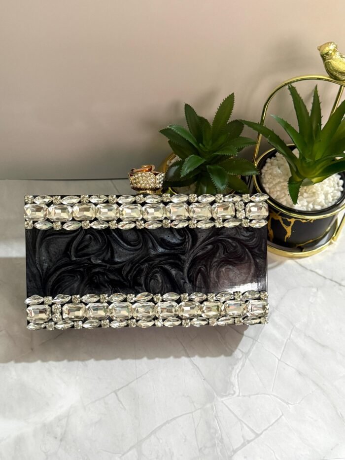 A black clutch adorned with a border of large clear crystals, placed next to decorative plants and a ring.