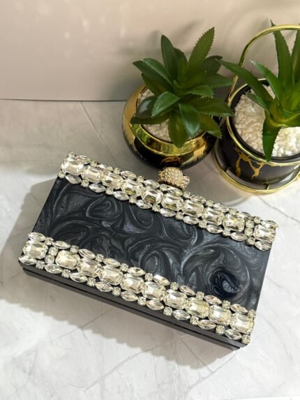 A black clutch adorned with a border of large clear crystals, placed next to decorative plants and a ring.