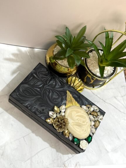 A black clutch with a central mother-of-pearl design, adorned with gold-tone leaves, floral accents, and sparkling crystals, placed next to decorative plants.