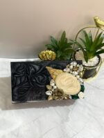 A black clutch with a central mother-of-pearl design, adorned with gold-tone leaves, floral accents, and sparkling crystals, placed next to decorative plants.