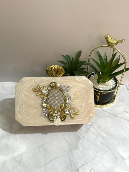 An ivory clutch with a crystal mesh center, surrounded by gold-toned leaves, pearls, and floral embellishments, placed next to decorative plants.