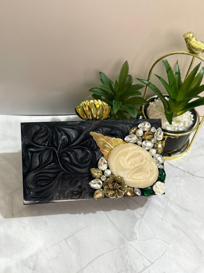 A black clutch with a central mother-of-pearl design, adorned with gold-tone leaves, floral accents, and sparkling crystals, placed next to decorative plants.