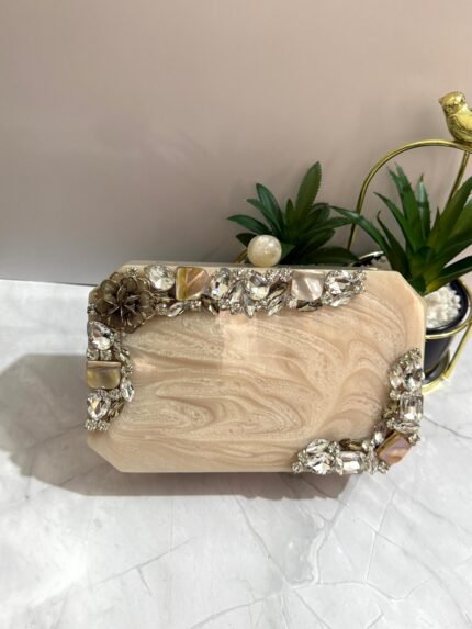 An ivory clutch with a crystal and mother-of-pearl embellished design, featuring floral accents and sparkling gems, placed next to decorative plants.