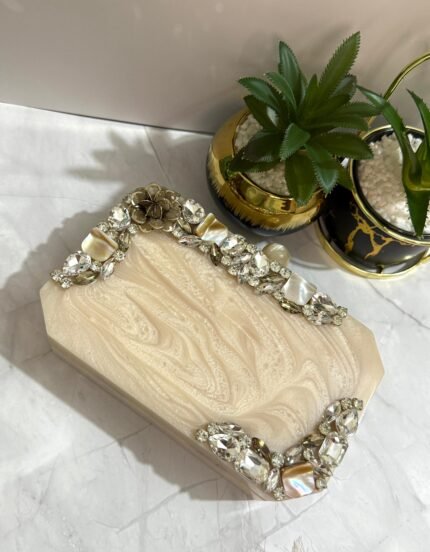 An ivory clutch with a crystal and mother-of-pearl embellished design, featuring floral accents and sparkling gems, placed next to decorative plants.