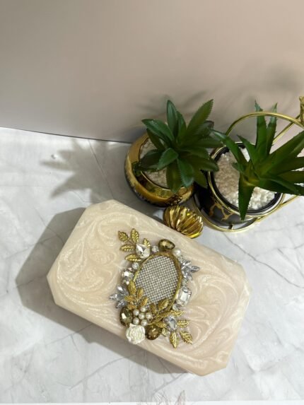 An ivory clutch with a crystal mesh center, surrounded by gold-toned leaves, pearls, and floral embellishments, placed next to decorative plants.