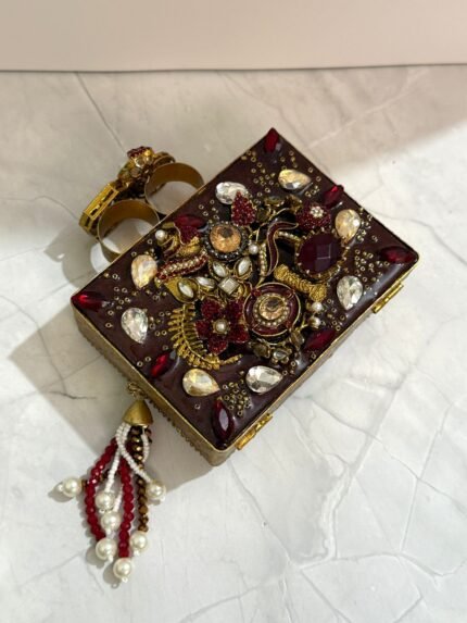 A luxurious red and gold embellished clutch with gemstones, pearls, and a pearl tassel, with two rings attached on top, placed on a marble surface.