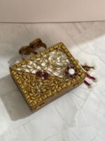 A luxurious red and gold embellished clutch with gemstones, pearls, and a pearl tassel, with two rings attached on top, placed on a marble surface.