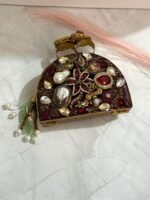 A red and gold embellished clutch with sparkling gemstones, pearls, and a pearl tassel, featuring intricate detailing, placed on a marble surface.