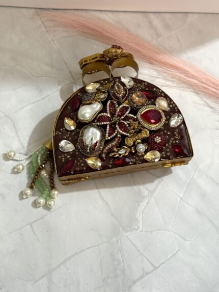 A red and gold embellished clutch with sparkling gemstones, pearls, and a pearl tassel, featuring intricate detailing, placed on a marble surface.