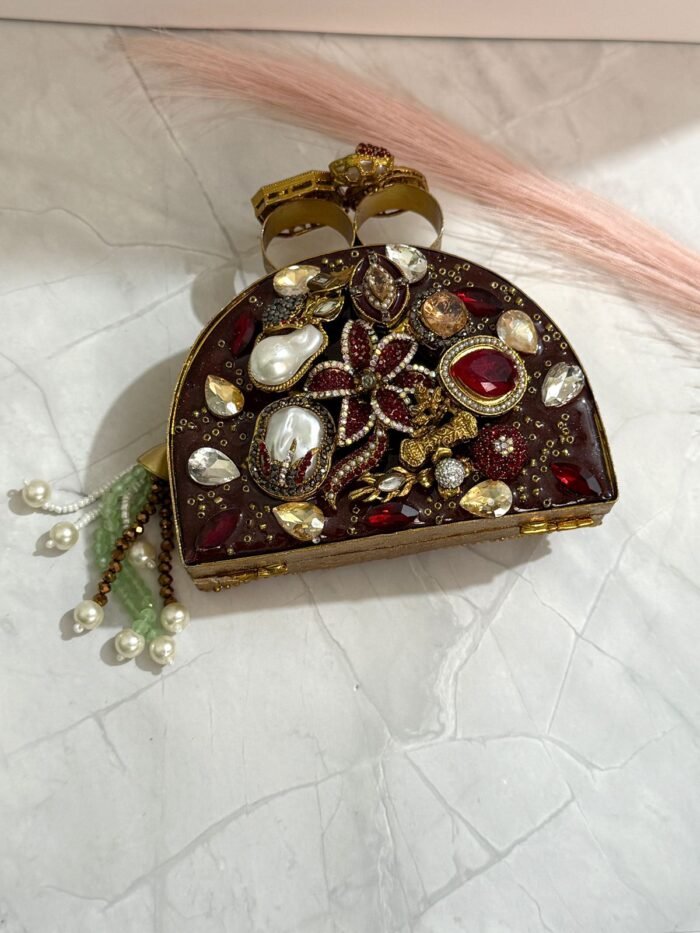 A red and gold embellished clutch with sparkling gemstones, pearls, and a pearl tassel, featuring intricate detailing, placed on a marble surface.