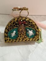 A red and gold embellished clutch with sparkling gemstones, pearls, and a pearl tassel, featuring intricate detailing, placed on a marble surface.