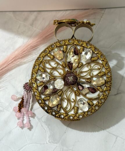 A gold clutch with crystal embellishments, featuring a floral design and pink bead tassel, placed on a marble surface.