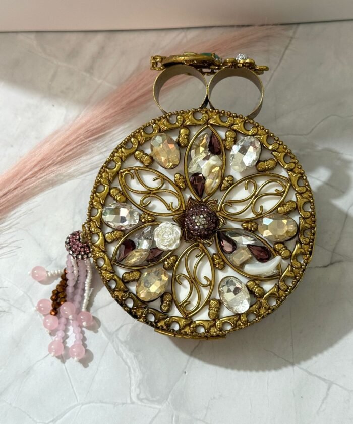 A gold clutch with crystal embellishments, featuring a floral design and pink bead tassel, placed on a marble surface.