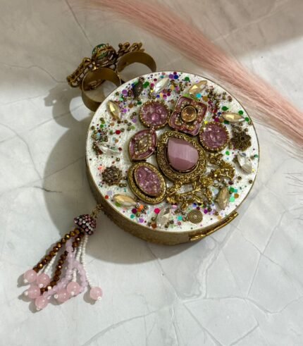 A gold clutch with crystal embellishments, featuring a floral design and pink bead tassel, placed on a marble surface.