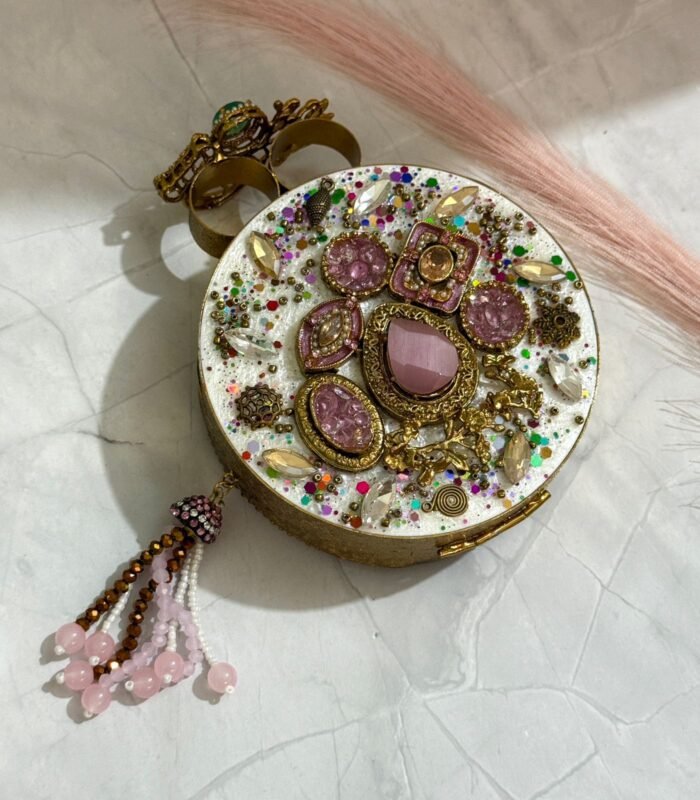 A gold clutch with crystal embellishments, featuring a floral design and pink bead tassel, placed on a marble surface.