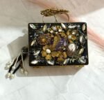 A black clutch with gold floral embellishments, large crystals, pearls, and a pearl tassel, placed on a marble surface.