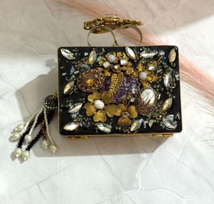 A black clutch with gold floral embellishments, large crystals, pearls, and a pearl tassel, placed on a marble surface.