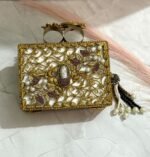 A black clutch with gold floral embellishments, large crystals, pearls, and a pearl tassel, placed on a marble surface.