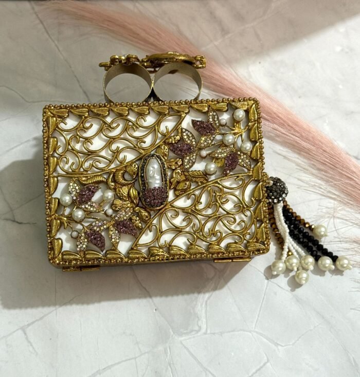 A black clutch with gold floral embellishments, large crystals, pearls, and a pearl tassel, placed on a marble surface.