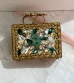 A gold clutch with crystal embellishments and a green stone centerpiece, featuring emerald and clear gemstones, placed on a marble surface.