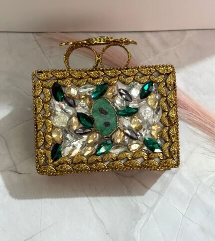 A gold clutch with crystal embellishments and a green stone centerpiece, featuring emerald and clear gemstones, placed on a marble surface.