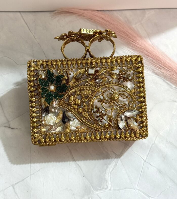 A gold clutch with crystal embellishments and a green stone centerpiece, featuring emerald and clear gemstones, placed on a marble surface.