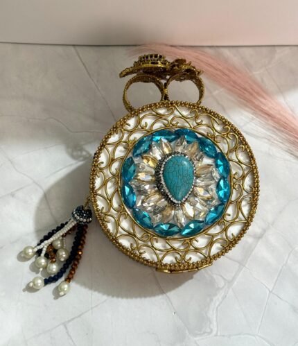 A turquoise and gold clutch adorned with sparkling crystals and a pearl tassel, placed on a marble surface.