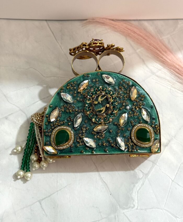 A green clutch with gold accents, sparkling crystals, green gemstones, and pearl tassels, placed on a marble surface.