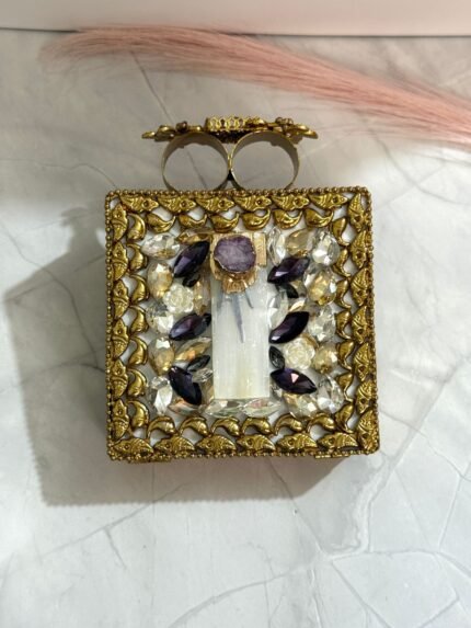A gold floral and crystal embellished clutch with lavender gemstones, featuring intricate detailing, placed on a marble surface.
