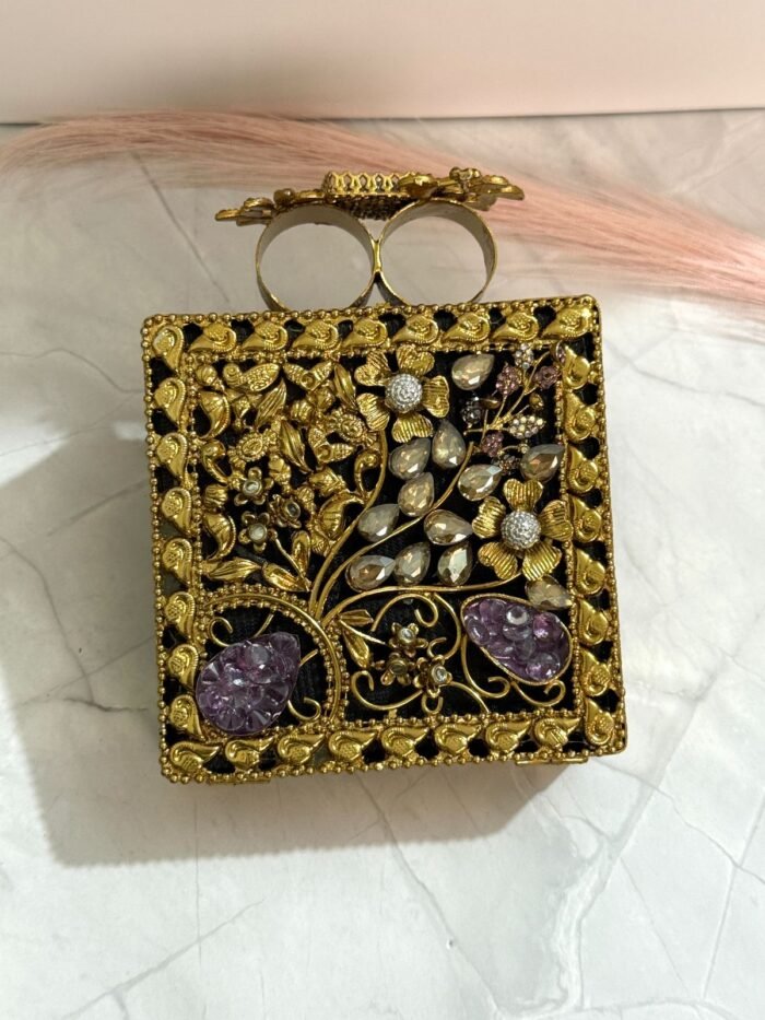 A gold floral and crystal embellished clutch with lavender gemstones, featuring intricate detailing, placed on a marble surface.