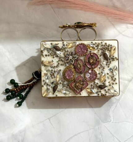 A pink and gold embellished clutch with crystal and gemstone detailing, featuring a beaded tassel, placed on a marble surface.