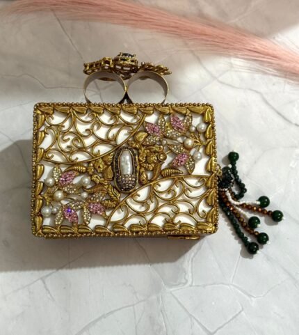 A pink and gold embellished clutch with crystal and gemstone detailing, featuring a beaded tassel, placed on a marble surface.