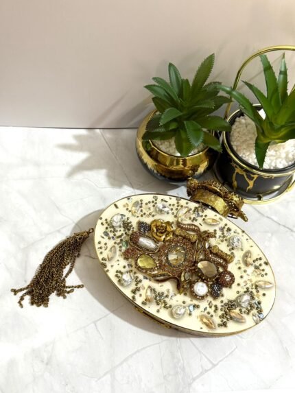 A gold and crystal embellished clutch with pearls, a rose accent, and a tassel detail, placed alongside decorative plants.