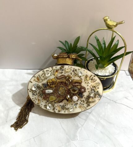 A gold and crystal embellished clutch with pearls, a rose accent, and a tassel detail, placed alongside decorative plants.