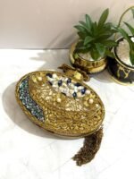 A gold and crystal embellished clutch with pearls, a rose accent, and a tassel detail, placed alongside decorative plants.