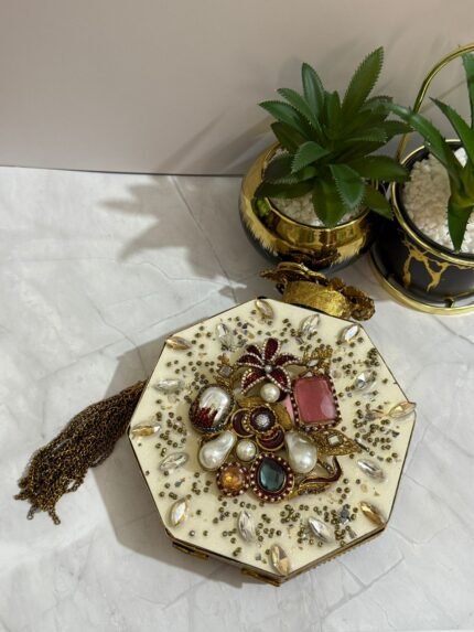 A luxurious octagonal clutch with gold accents, jewel-toned crystals, pearls, and a tassel detail, placed alongside decorative plants.