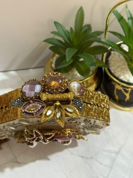 A luxurious octagonal clutch with gold accents, jewel-toned crystals, pearls, and a tassel detail, placed alongside decorative plants.