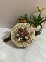 A luxurious octagonal clutch with gold accents, jewel-toned crystals, pearls, and a tassel detail, placed alongside decorative plants.